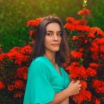 Woman in a teal dress posing amidst vibrant red flowers in a garden, Bauru, Brazil.